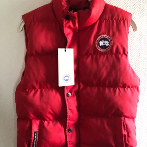 Röd dunväst från Canada Goose - Säljer en snygg röd dunväst från Canada Goose med deras ikoniska logga på bröstet. Västen har en hög krage och knäppning med både dragkedja och knappar. 
