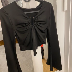 Svart croppad topp med knyt och utsvängda ärmar - Trendig svart croppad topp med långa utsvängda ärmar och snygg knytdetalj framtill. Toppen har en lätt rynkad effekt vid bysten och en v-ringad halsringning. Perfekt för att lyfta vilken outfit som helst.
