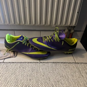Nike Mercurial fotbollsskor lila och gula OG - Säljer ett par Nike Mercurial fotbollsskor i lila och gult med snörning och rund tå. Skorna har en snygg kontrast mellan de två färgerna och är perfekta för dig som vill sticka ut på planen.