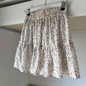 Blommig volangkjol från SHEIN - Supersöt kort blommig kjol från SHEIN med volanger och smock i midjan. Kjolen är vit med små blommor i orange, blått och svart. Perfekt för en fräsch och somrig look!