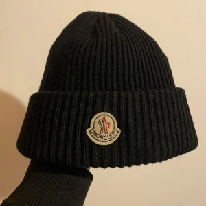 Svart ribbad mössa från Moncler - Snygg svart ribbad mössa från Moncler med deras ikoniska logotyp framtill. Perfekt för att hålla värmen under kyliga dagar.