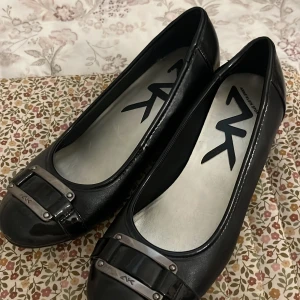 Svarta ballerinaskor med metallspänne från AK - Säljer ett par svarta ballerinaskor från AK med rund tå och dekorativt metallspänne framtill. Skorna har en stilren design och är tillverkade i syntetmaterial med glansig finish.