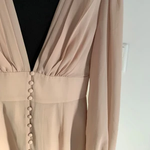 Beige långärmad klänning med knappar från NA-KD - Aldrig använt Säljer en elegant beige klänning från NA-KD i storlek 34. Klänningen har djup v-ringning, långa ärmar och dekorativa knappar framtill. Perfekt för dig som gillar en stilren och feminin look.