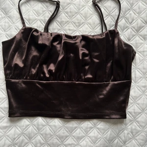 Brun satinlinne - Snygg brun satinlinne med smala axelband och en croppad passform. Perfekt för en stilren look. Axelbanden är justerbara för bästa passform.