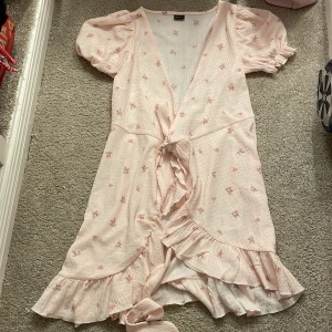 Blommig klänning från Gina Tricot - Söt blommig klänning från Gina Tricot i ljusrosa med små blommor. Klänningen har korta puffärmar och volangdetaljer framtill samt en knytning i midjan. Perfekt för varma dagar. använt en gång jättefin klänning men har fått nya så kommer jag inte till användning