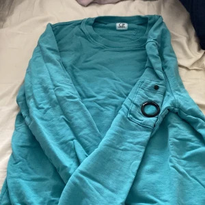 Turkos sweatshirt från C.P. Company - Säljer en turkos sweatshirt från C.P. Company i mjuk bomull. Tröjan har lång ärm och en unik detalj med en svart ring på ena ärmen. Perfekt för dig som gillar streetwear och vill sticka ut lite extra. Cp company loggan försvann i tvätten 