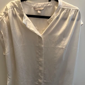 Vit blus från H&M - Säljer en stilren vit blus från H&M i storlek L. Blusen har korta ärmar, v-ringad hals och är tillverkad i ett mjukt, glansigt material som ger en elegant känsla. Perfekt att matcha med jeans eller kjol för en clean look. Blusen är endast använd fåtal gånger och är i princip i nyskick. 🤍