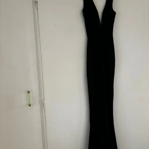 BALKLÄNNING Svart långklänning med djup v-ringning - Säljer en elegant svart långklänning med djup v-ringning och smala axelband. Klänningen har en figurnära passform och är perfekt för festliga tillfällen. Enkel och stilren design som ger en snygg siluett.