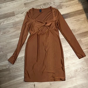 Brun klänning från Shein - Snygg brun klänning från Shein i en mjuk och glansig polyester. Klänningen har en v-ringad design med långärmade ärmar och en knytning framtill. Perfekt för en stilren look! Liten defekt under högra armen. 
