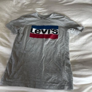 Grå t-shirt från Levi's - Säljer en klassisk grå t-shirt från Levi's med deras ikoniska logga i blått, svart och rött på bröstet. Perfekt för en avslappnad stil. Passar till det mesta och är ett måste i garderoben!