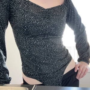 Svart/mörkgrön mönstrad omlott bodysuit - Säljer en Mörkgrön nästan svart bodysuit med vitt prickigt mönster och omlott-detalj framtill. Toppen har långa ärmar, v-ringning och knäppning i grenen. Snygg och figurnära passform som framhäver midjan. Den är använd 1 gång med trosor såklart och knäpps mellan benen. 