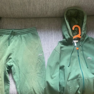 Grön dress från Lacoste - Säljer en grön dress från Lacoste med dragkedja och huva. Tröjan har en broderad logotyp på bröstet och snörning vid huvan. Perfekt för en avslappnad stil.