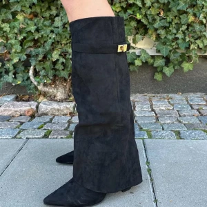 Svarta höga stövlar med spänne - Snygga svarta höga stövlar i mocka med dekorativt guldfärgat spänne upptill. Modellen har spetsig tå och en stilren design som passar perfekt till höst- och vinterlooken.