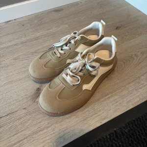 Beige och olivgröna sneakers med vita detaljer - Snygga sneakers i beige och olivgrönt med vita detaljer och snörning. Skorna har en rund tå och en kombination av mocka och syntetmaterial. Perfekta för en trendig vardagslook.
