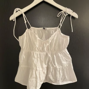 Vit linnetopp från H&M Divided - Säljer en söt vit linnetopp från H&M Divided med tunna axelband och volangdetaljer. Toppen har en luftig passform och är perfekt för varma sommardagar. Den är tillverkad i ett lätt och bekvämt material.