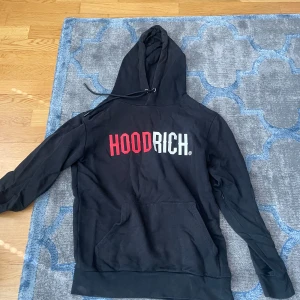 Svart hoodie från Hoodrich med tryck - Svart hoodie från Hoodrich med stor logga i rött och vitt framtill. Tröjan har huva med dragsko, magficka och en dold ficka med dragkedja på ärmen. Perfekt för en streetwear-look.