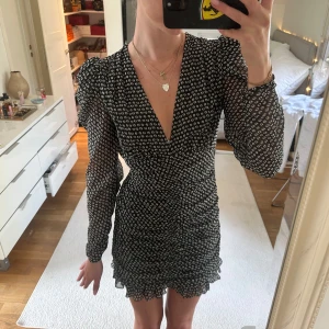 Svart kortklänning med vita mönster och volang - Säljer en svart kortklänning från Zara med små vita mönster, v-ringning och långa puffärmar. Klänningen har volangdetaljer nertill och är figurnära med lätt rynk vid midjan. 