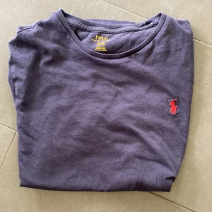 Mörkblå t-shirt från Polo Ralph Lauren - Säljer en mörkblå t-shirt från Polo Ralph Lauren med klassisk röd broderad logga på bröstet. T-shirten har rund halsringning och korta ärmar. Perfekt för en stilren och avslappnad look.