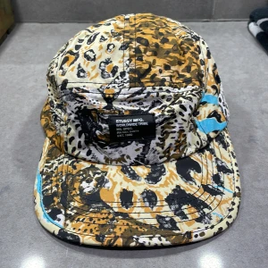 Leopardmönstrad keps från Stussy - Snygg keps från Stussy med leopardmönster i svart, beige och brunt. Kepsen har en justerbar rem baktill och en etikett framtill med Stussy-loggan. Perfekt för att ge din outfit en cool touch.