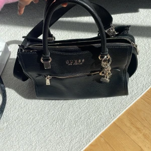 Guess Väska - Äkta Guess väska i färgen svart med axelband. Helt ny och aldrig använd, kommer i dustbag. En stor ficka och två lite mindre på sidorna av den stora fickan. 