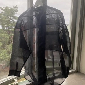 Svart genomskinlig skjorta, Monki strl M - Jättefin svart skjorta från Monki, i organzamaterial. Genomskinlig och har lite oversize passform. Perfekt till nyår! Storlek Medium. 