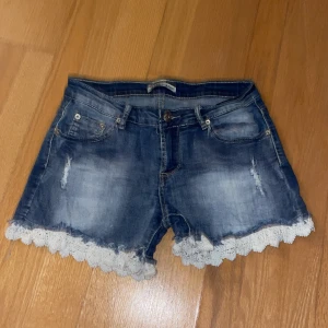Jeansshorts med vita volanger - Två slitningar på sidorna och små slitningar på bakfickorna (köptes så). Vita volanger med blommönster längst ner på shortsen. Två framfickor och två bakfickor  Väldigt bra skick 