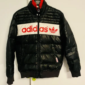 Adidas dunjacka stl S - Svart puffer jacka Adidas med stort tryck i vintage stil, märkt storlek S men passar även M. Många snygga detaljer, se bilder för skick. Inga synliga skador vad jag kan hitta.   Bröstvidd: ca 108 cm