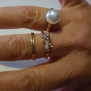Tre guldfärgade ringar. En ring med roset och vita stenar storlek 18. En slät ring storlek 18 och ringen med en vit pärla Storlek justerbar från storlek 18. Alla tre för 15kr och 1 för 5kr.