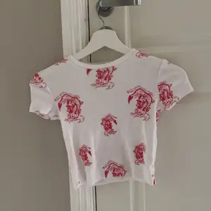 En kort vit t-shirt/topp med röda drakar på i ribbat material. Plagget är endast provat och därför i nyskick. Den är från berskha i storlek XS. Jag kan leverera varan om köparen står för leveranskostnaderna😊