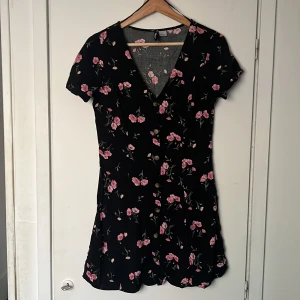 Blommig klänning - Kort svart klänning med blommor. Knappar längs med. Knappt använd. Strl 40.