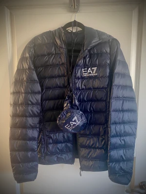 Emporio Armani dunjacka EA7 storlek XS - Emporio Armani EA7 dunjacka i mörk marinblå med luva. Använd ett fåtal gånger, ser helt ny ut. Kommer med förvaringspåse. Djur- o rökfritt hem. Perfekt till våren!