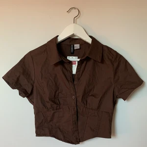 Brun korsett/skjort topp - Superfin croppad brun topp från HM. Köpt på rea men tyvärr fel storlek, enbart testad 1 gång! Storlek 36, men mer lik 34.  Hoppas någon annan kan få glädje av den, verkligen supersnygg topp!!! Frakt tillkommer, men vid flera köp kan samtliga gå i 1 paket!