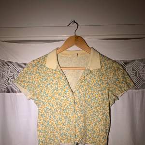 Somrig croppad skjorta i bomull från Urban Outfitters, med gulgrönt blommigt mönster. Använd endast ett fåtal gånger och i mycket bra skick. 