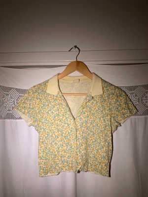 Croppad skjorta från Urban Outfitters  - Somrig croppad skjorta i bomull från Urban Outfitters, med gulgrönt blommigt mönster. Använd endast ett fåtal gånger och i mycket bra skick. 