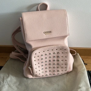 Guess ryggsäck - Rosa Guess ryggsäck, lite sliten baktill men går att använda om man ej har tunga saker i den. 30cm hög, 28cm bred och 12 cm djup.