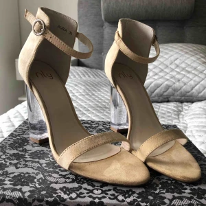  - Snygga klackar från Nelly. Beige mockaimitation, glasklar klack och spänne vid ankeln. Klackhöjd 10 cm. Aldrig använda! 