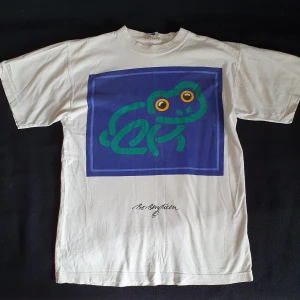 Vit t-shirt med tryck - Vit t-shirt med tryck av en groda i storlek L. Har ett litet lagat hål nedtill fram samt några dova ljusbruna fläckar, främst inuti nedtill som knappt syns (se bild 2). Fraktkostand blir 47kr.