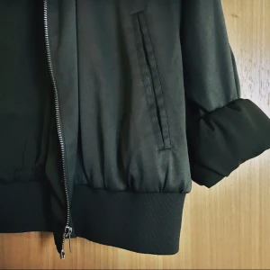  - Tunn jacka i bomberstil. Militärgrönt, glansigt material.  Sparsamt använd, gott skick. Passar S-L, beroende på passform. Märkt storlek L. 