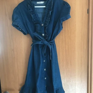 Söt och somrig Jeansklänning - En väldigt söt jeansklänning. Märken från där skärpfållarna brukade sitta men syns inte så mycket. Speciellt inte med skärpet runt midjan. Fina volangdetaljer. Skulle passa någon som är 40-42 och mindre byst. 99% bomull 1% elastan