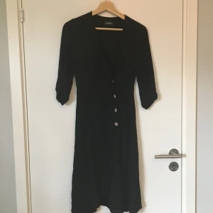 Svart klänning med omlottknäppning Monki - Svart klänning med omlottknäppning från Monki. Tror det är storlek XXS men kan också vara XS, lappen saknas. Gott skick. Frakt betalas av köparen.