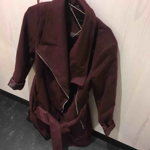  - Jättefin kappa i vinröd färg med snygga detaljer!! Knappt använd. Med skärp och dragkedja, modellen kan varieras i utseende beroende på hur man stänger den.  två fickor på sidan. Perfekta höstjackan! 