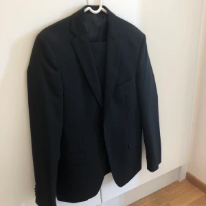 Svart kostym - Svart kostym (både kavaj och byxor) från Andersson Herr. Storlek 48. Material: 44% ull, 54% polyester, 2% eldarna.