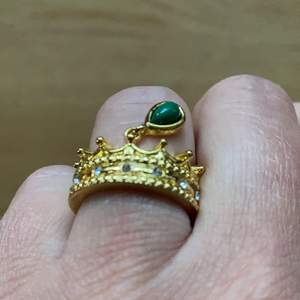 Otroligt söt helt ny ring i guldfärg ca 16,7 mm. Är formad som en krona med vita gnistrande stenar och en grön hängande pärla. 
