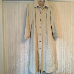 Beige vintage trenchcoat lång M - En supersnygg vintage trenchcoat i beige. Fodrad i beiget satintyg. Ger en jättefin silhuett och går in lite i midjan. Fin krage och knappar!