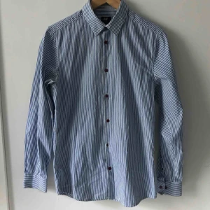  - Blåvit-randig skjorta, har en mindre reva vid en knapp
