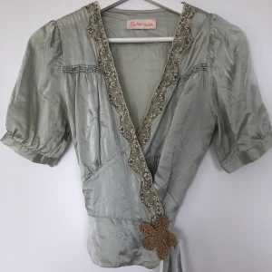 Stella Nova Silkes blus  - Har en liten skada på ena axeln, märks knappt (se bild) annars är den i bra skick!  Guldiga blomman går att sprätta upp ifall man ej vill ha den.  Färgen är ”silver grön”. Storlek står ej men den passar som en XS/S.  44kr för frakt/träffas i Kista.