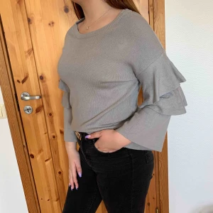  - Grå tjockare tröja med volangärmar, passar en M, kortare i ärmen. Köparen står för frakten