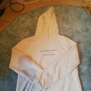 Hoodie - En ljusrosa hoodie från gina med trycket DU ER IKKE ALENE vilket är ett citat från den norska serien SKAM. En liten ljus fläck finns på vänster arm men annars är den ok. 