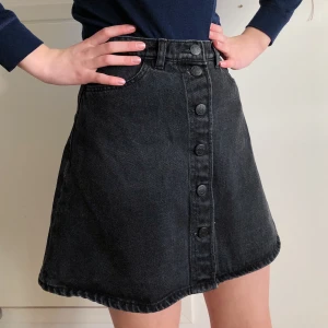 Svart jeanskjol - Kjol från Monki. I svart denim med knappar längs framsidan. Relativt högmidjad. Klockad i passformen. Strl: 36. CN: 165/68A. Längd: 42 cm. Vikt: 365 g. Samfraktar gärna! 