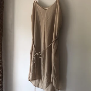 Beige klänning - Beige klänning med spagetti straps. Knytning i midjan. Längre bak. Undrerklänning i trikå. Från Phanuel. Inköpt i USA, stormed framgår ej men passar S! Köparen står för frakt.